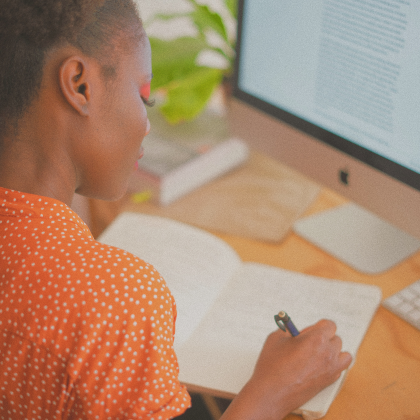 Girl writing in notebook and working on Apple desktop.
